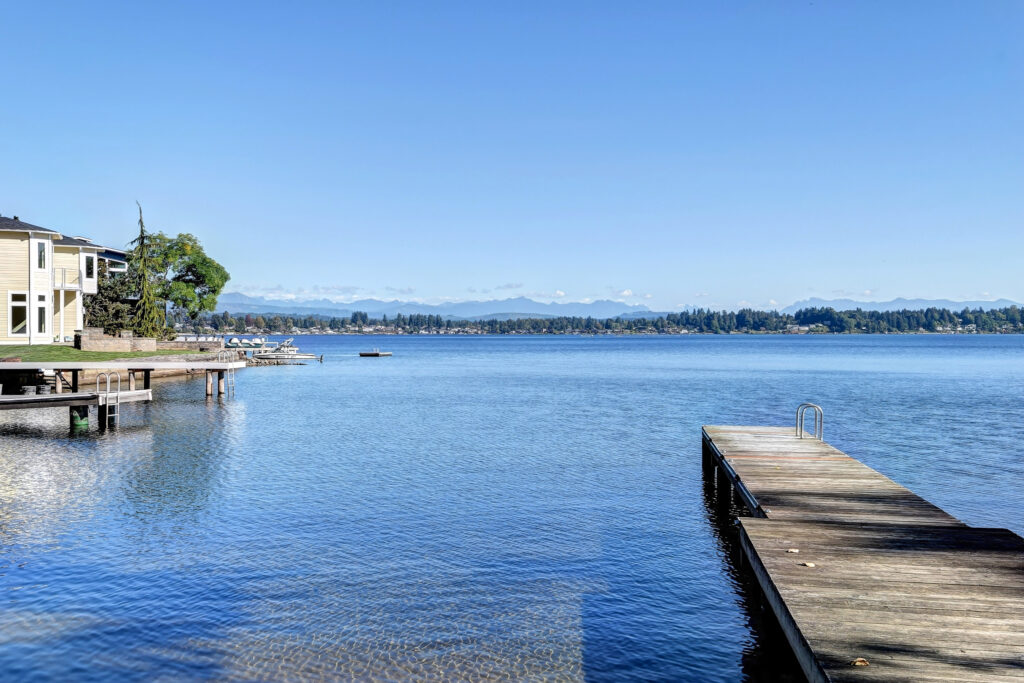 A picture of a dock on Lake Stevens with some homes and a view of the trees and mountains in the distance. Beautiful blue water and sky.